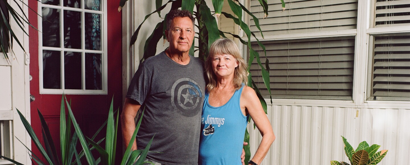 A man and woman with blonde hair stand next to each other outside their home with plants in the background and a red door.