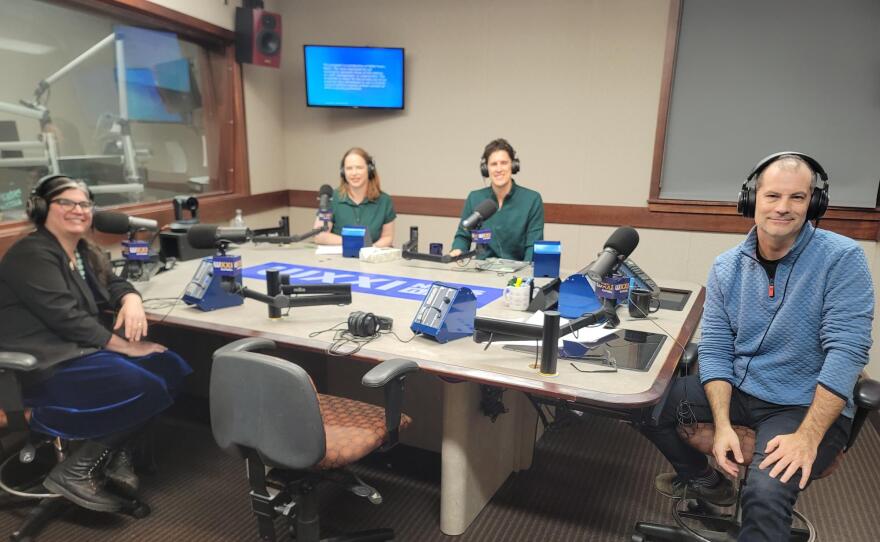 Four smiling people wearing headphones sit at a table in a radio talk studio: a woman front left has long dark hair and is wearing glasses, a black blazer, a blue velvet skirt and black boots, a man front right has very short dark hair and is wearing a light-blue quilted pullover, jeans and sneakers; a woman back left has shoulder-length red hair and is wearing a short-sleeved green blouse; a man back right has short dark hair and is wearing a dark green button-down shirt.