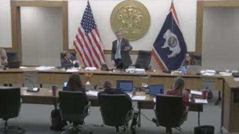 Five people sit at a long desk. The man in the center is standing and gesturing with his left hand.