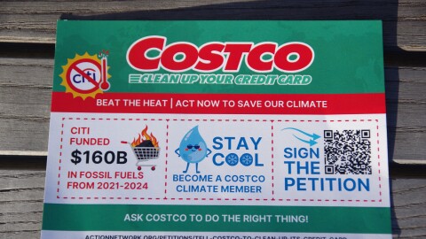 A small rectangular flyer sits on a wooden bench, zoomed in close to the small advertisement. The flyer promotes a petition against CostCo, for their partnership with Citibank, citing billions of dollars Citibank sent to the fossil fuel industry. 