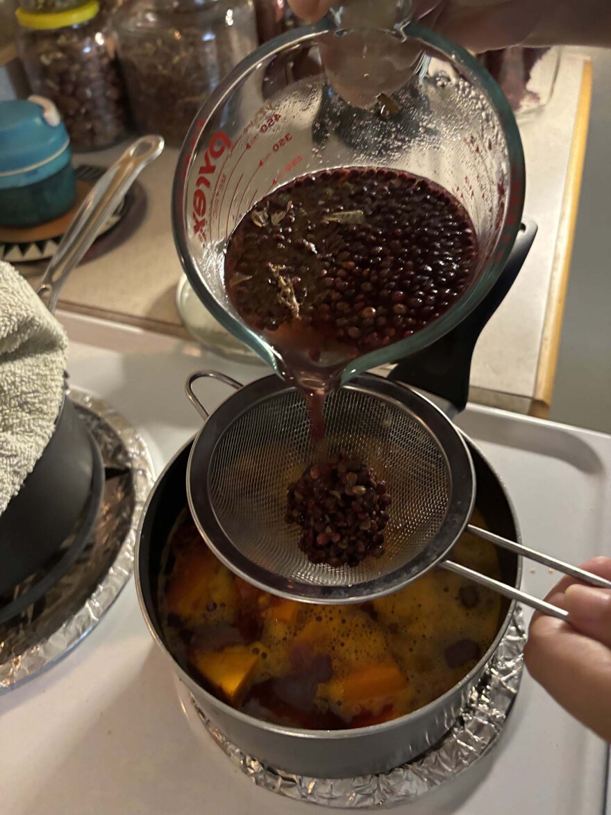 measuring cup with dark liquid and seeds pouring liqid into hand held strainer over a pot with orange chunks of squash