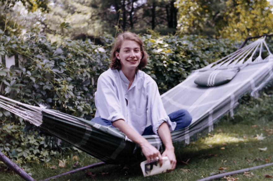 A young Mary Oliver sits criss-cross in the center of a green plaid hammock, facing over the side. Her hands hang off the hammock, holding a book. The hammock is in a space with green grass and hedges behind.