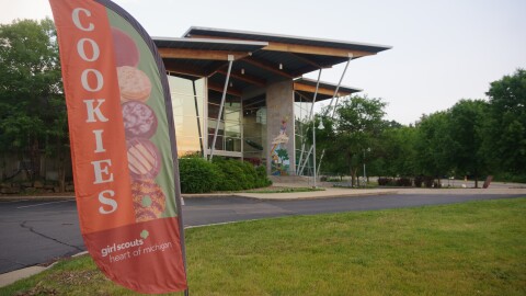 A photo of the Girl Scout Heart of Michigan, Kalamazoo Regional Center on June 17, 2024.  In the foreground is a flag style lawn banner that reads "cookies."  The word is spelled out in white letters on a light orange background with the letters stacked on top of each other vertically on the left side of the banner. On the right vertical side of the banner on a green background is a picture of five types of Girl Scout Cookies, stacked from the top to the bottom of the flag.  And at the bottom, in the lower quarter of the banner in white letters on a dark orange background it reads "Girl Scouts Heart of Michigan" in all lowercase letters.   Behind the flag is the driveway around the front of the concrete building with large panes of glass and a colorful mural on the concrete wall.  The building has  three distinct roof lines that are designed to loosely resemble a person with arms outstretched.  Bushes and trees are close to the building, on the building side of the drive.  A yellow-green lawn is in the foreground.
