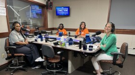 Four smiling people wearing headphones sit around a table in a radio talk studio: A woman front left has long black hair and is wearing a gray polo shirt black pants, and white sneakers; a man back left is wearing glasses, a black hat, and an orange t-shirt; a woman back center has long dark hair and is wearing an orange t-shirt; a woman at right has shoulder-length blonde hair and is wearing a green collared shirt, white pants, and white shoes
