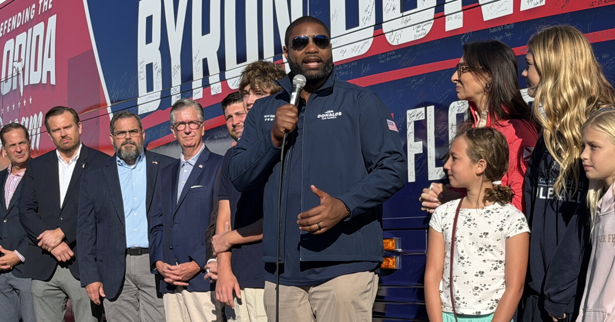 Photo: wusf.org A man in blue jacket and dark sunglasses stands before a bus bearing his name, flanked by politicians, his wife, and also