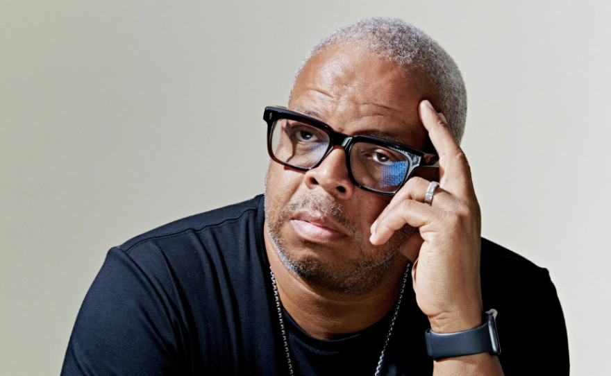 A man with very short grey hair is wearing glasses and a black long-sleeved shirt is sitting at a table holding a trumpet.