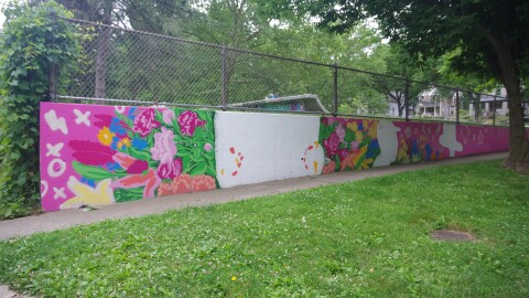 A mural is pictured on a cloudy day. The mural is on a long wall and is pink with many flowers. There are large blank spaces. 