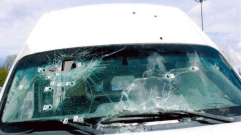 The front windshield of a white van, heavily cracked and pierced by multiple bullet holes with numbered police evidence markers placed next to the impact points.