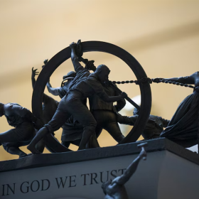 A close up of a mini model of people on top of a platform that reads "In God We Trust." The figures on top of the platform are pulling and pushing a hoop.