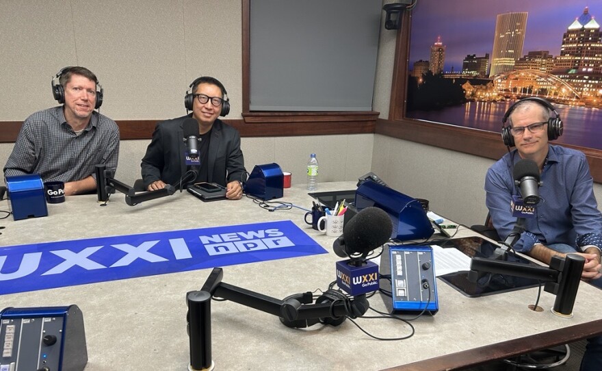 Three smiling men wearing headphones sit at a table in a radio talk studio: a man at left has short brown hair and is wearing a blue and white plaid button-down shirt; a man at center has short black hair and is wearing glasses and a black shirt under a dark grey suit coat; a man at right has very short dark hair and is wearing glasses and a blue button-down shirt
