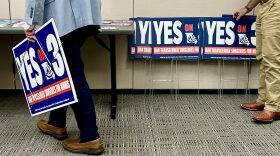 Two people from the waist down, one on either side of a conference room, are holding "Yes on 3" yard signs.