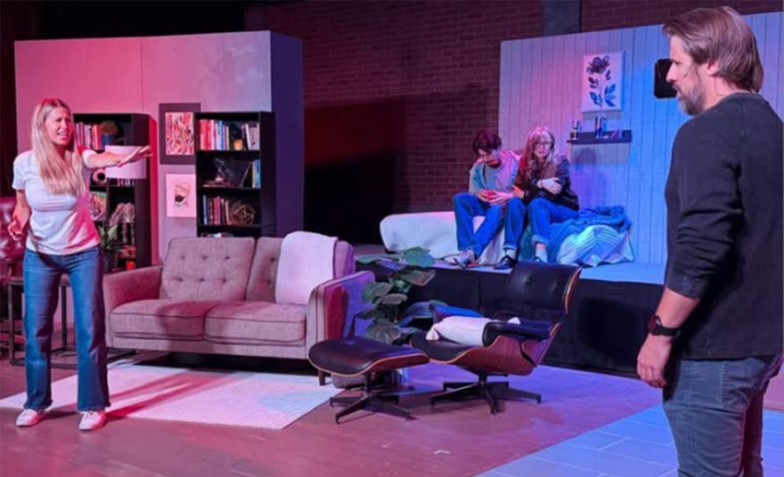 On a theatre stage, a woman with long blonde hair reaches out a hand toward a man with his back to the camera. Between them are two teenagers that sit next to each other on a bed. 