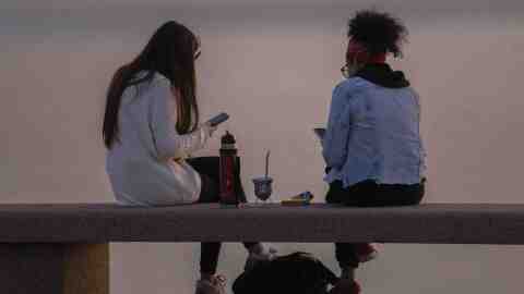 Two women sit on a bench at sunset, enjoying some snacks and looking at their phones