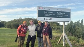 Four women stand beneath a sign that reads "York Factory: National Historic Site of Canada." Green grass and small bushes surround them. They are wearing wind breakers and two are wearing knitted hats. 
