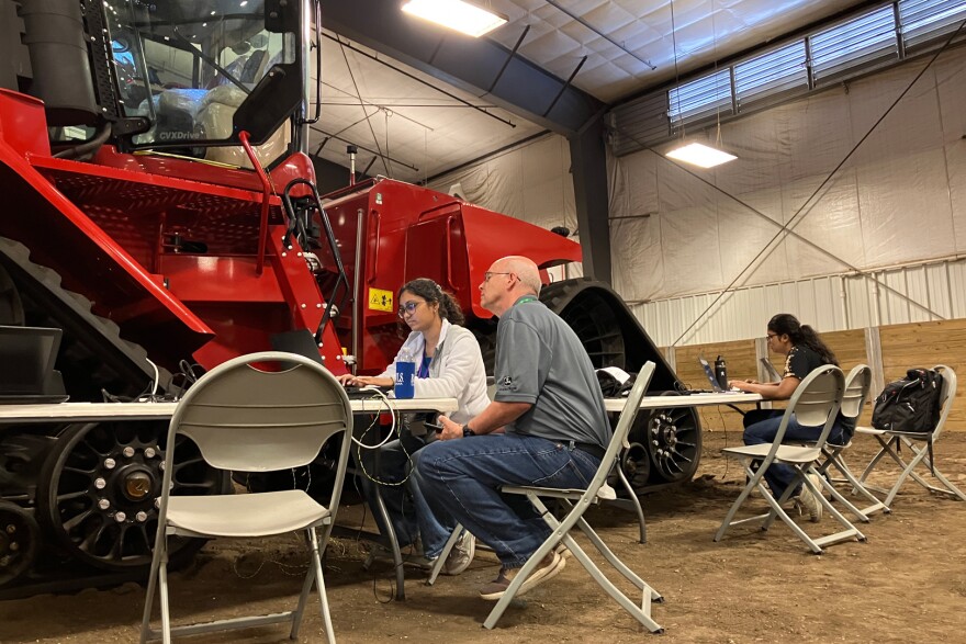 A woman with a pony tail types on a laptop next to a red tractor. A man sitting next to her looks at the screen.