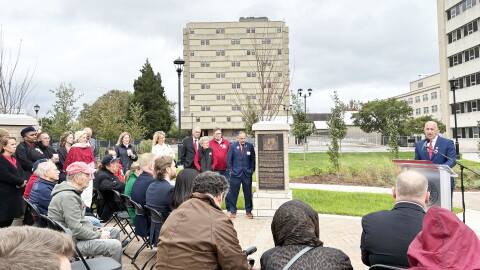 A man in a suit speaks at a podium during a dedication ceremony for a memorial plaque outdoors, with a seated audience watching.