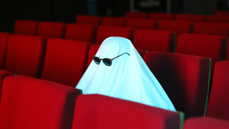 A sheet draped over a figure wearing sunglasses, sitting among red cinema seats, creating a humorous ghost-like appearance.