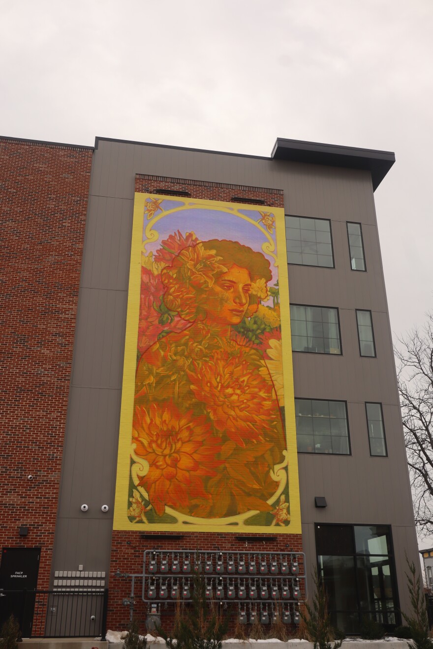 A mural by Colombian artist Natalia Gallego Sánchez, also known as Gleo, is seen on the side of Fulton Lofts in Cedar Rapids, Iowa. The mural had to be shipped, scanned and constructed without her after she was denied entry into the U.S. last summer. The large, vertical mural consists of mostly warm colors: oranges, yellows, reds. A woman's face can be seen at the top of the flowers. Her eyes are sad. Her gaze is to the side. The mural is surrounded by a bright yellow border. 