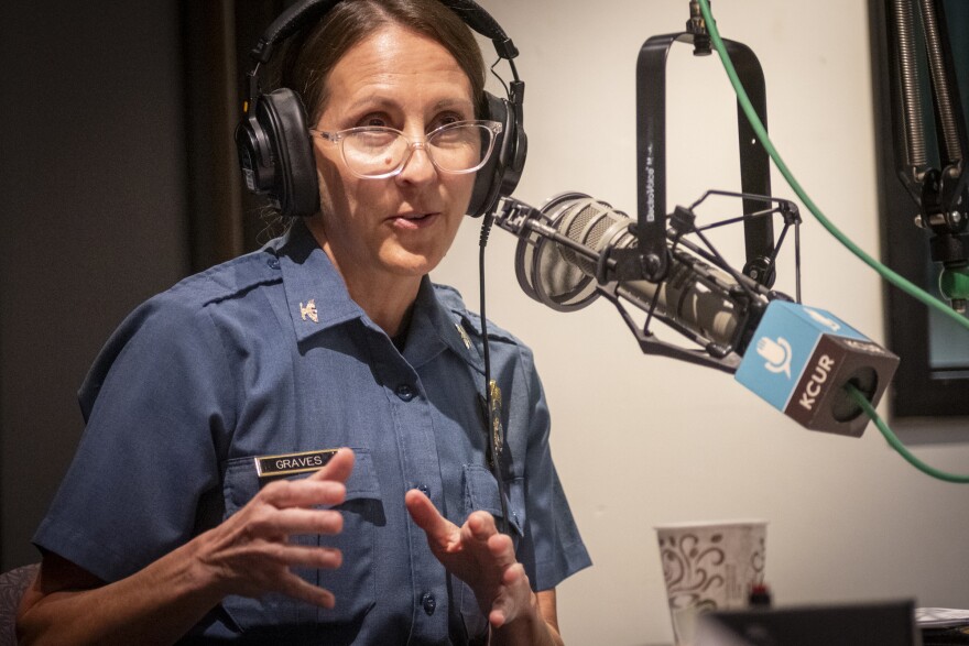 A person wearing a police uniform sits inside a radio studio. She is gesturing with both hands and talking at a microphone.