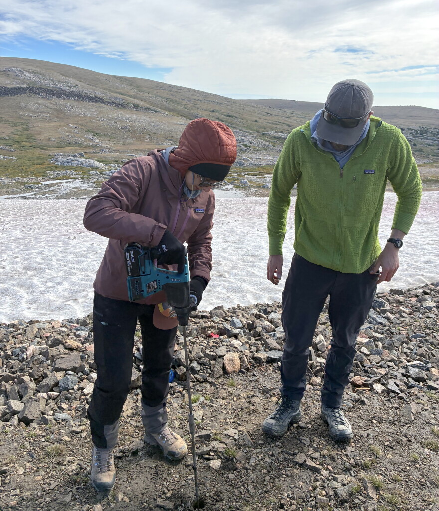 Two researchers use a power drill in rocky ground beside a high-elevation ice patch.