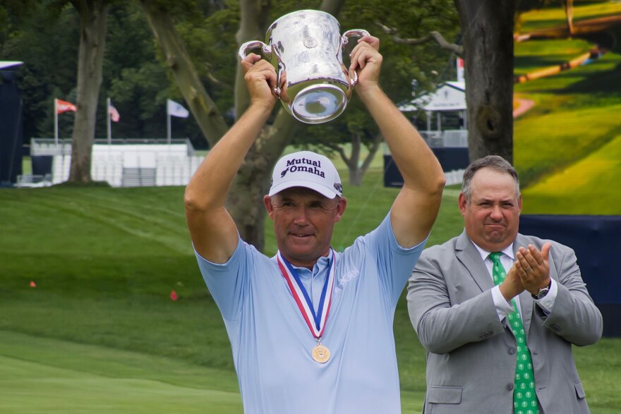 Senior golf player Padraig Harrington lifts up a trophy over his head while a tournament official looks on and claps.