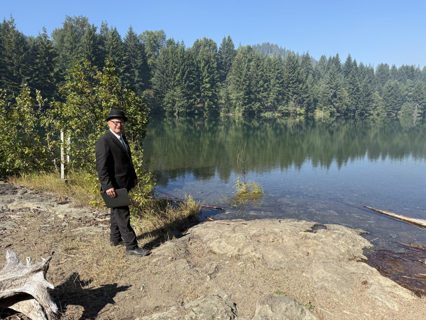 A man in a black suit and hat stands on the shore of a still lake, holding a document folder. 