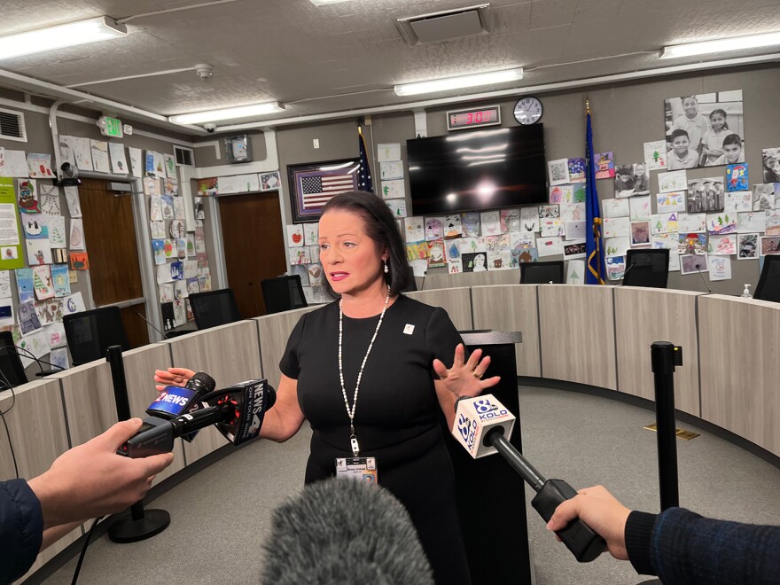 A woman in black stands in front of the dais in the Washoe County School District Board of Trustees meeting room and speaks toward several microphones. In the background, there are student-made holiday cards attached to the wall.