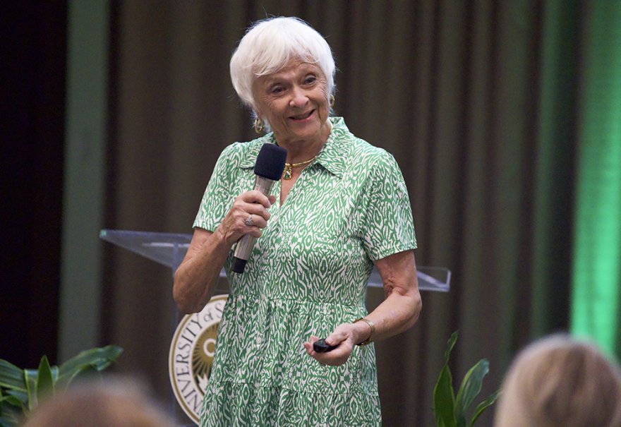 A woman with short white hair and wearing a green and white dress holds a microphone and speaks to an unpictured audience.
