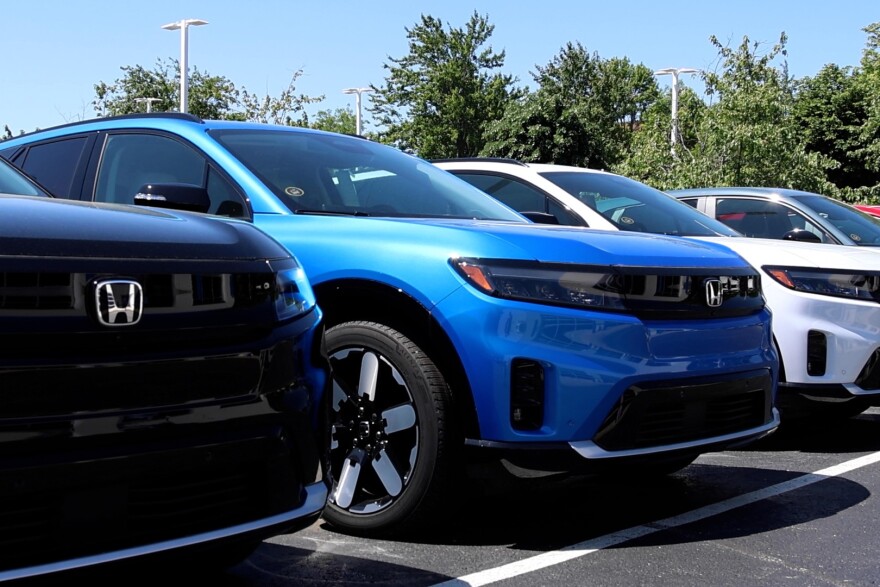 Honda electric vehicles lined up in the parking lot of a car dealership. There are 3 SUVs in black, blue and white. 