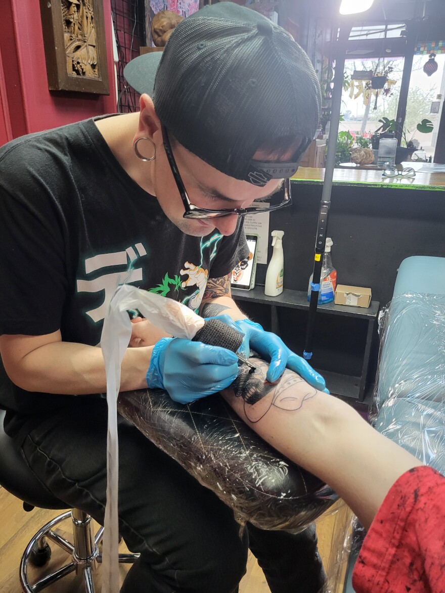 Jake Nolan wears blue latex gloves and holds a needle while he inks a design on Zach Erickson's arm