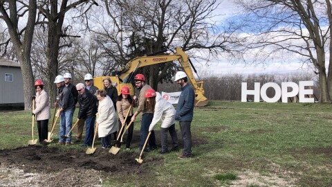A group of men and women push gold shovels into a pile of dirt in a grass field, with trees, a construction vehicle, and large white letters spelling "hope" in the background.  
