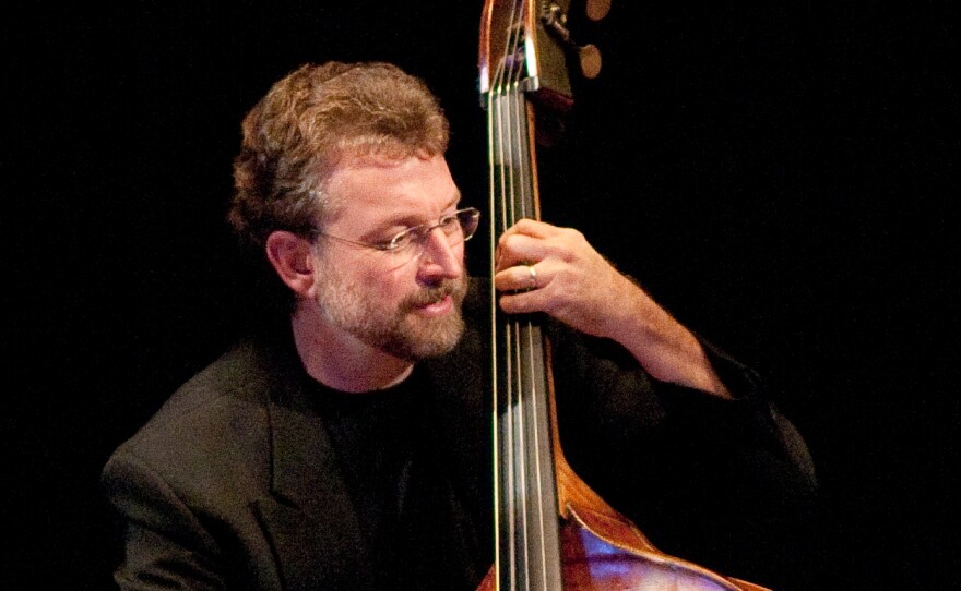 A man wearing glasses and dark clothing plays the bass in front of a black backdrop.