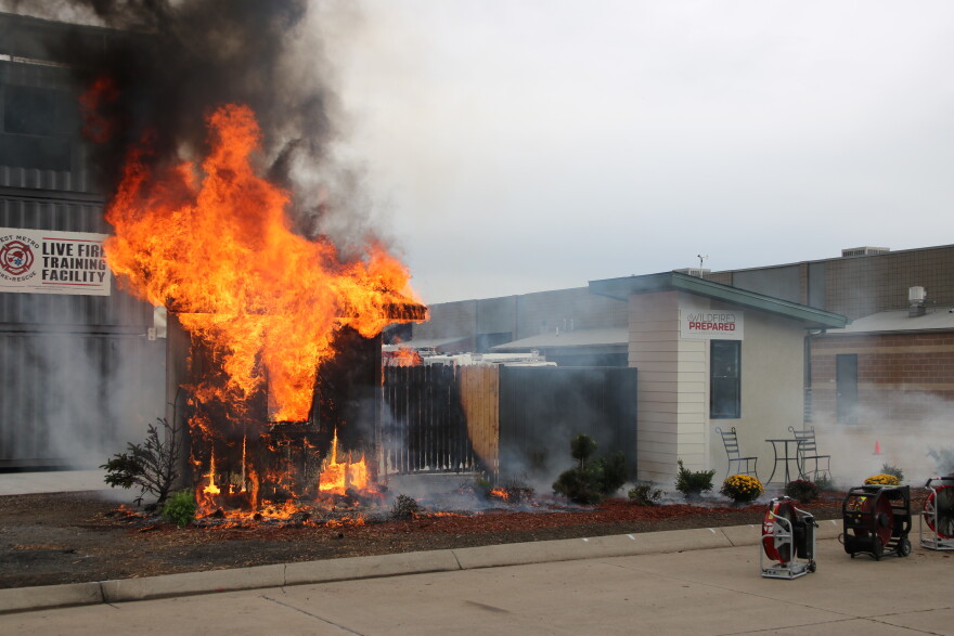 Two homes stand on a mulch lot. One is fully engulfed in flames and has thick black smoke rising from the roof. The wooden fence next to it is mostly singed. The home next to it has no signs of fire damage. It has a metal fence and a metal patio set.