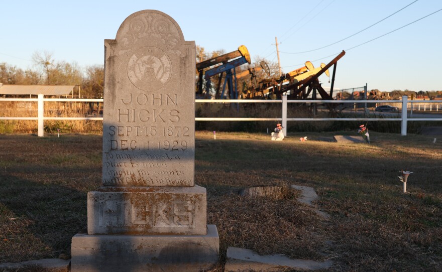 A headstone for John Hicks at the Oak Grove-Johnston Cemetery. Rusty pumpjacks loom in the background like rusty dinosaurs. 