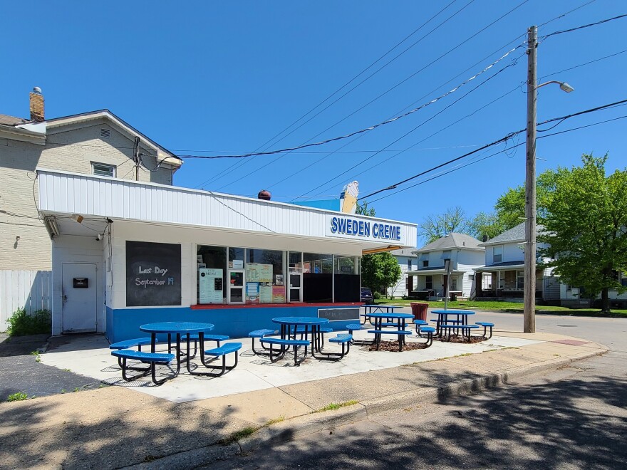 White building with blue lettering "Sweden Creme." Blue metal tables line a sidewalk. On top of the building is an ice cream cone sign.