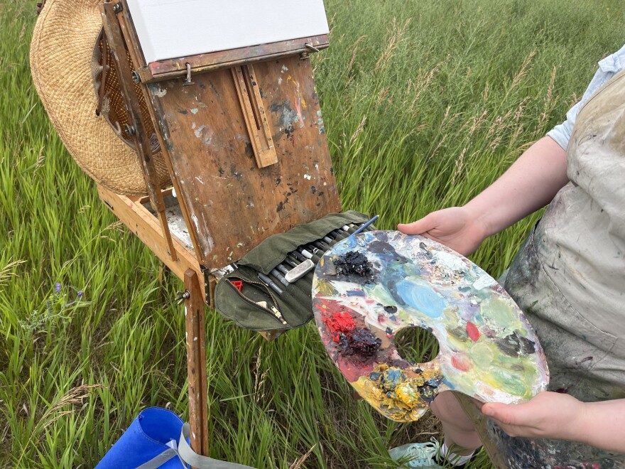 Artist's hands holding a palette splattered with paint in front of an easel. 