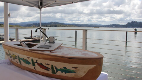 A small wooden canoe decorated with paintings of trees and fish on a display table set up on a boat ramp overlooking a bay. 