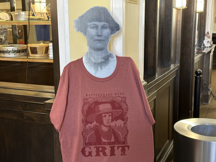 A black-and-white paper cutout of Rattlesnake Kate adorned with a red t-shirt that has an image of Rattlesnake Kate on it and the word "grit" underneath it.