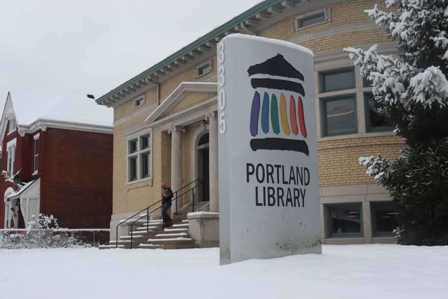 A snow-covered sign that says "Portland Library" at the front entrance of the snowy building