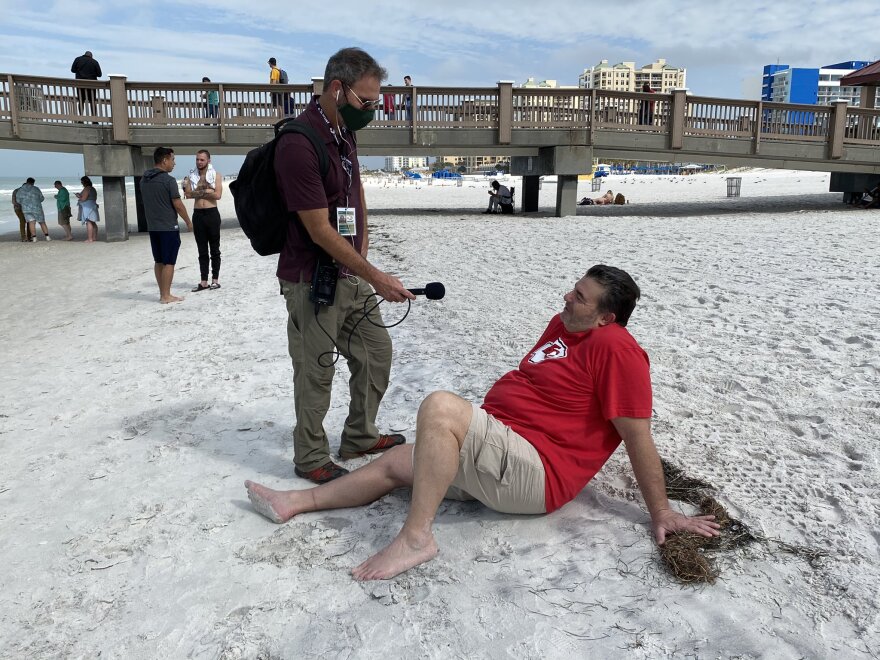 A reporter wearing a mask holds a mic out to a man in a red shirt sitting on the beack without a mask.