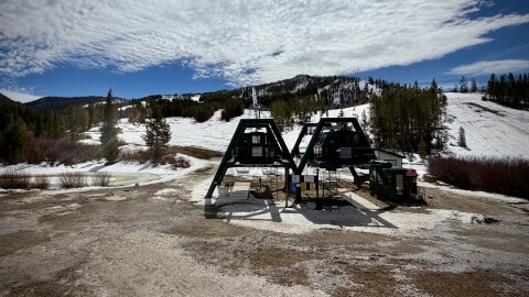 A ski lift surrounded by dirt and some snow further up the mountain on a mostly sunny day.