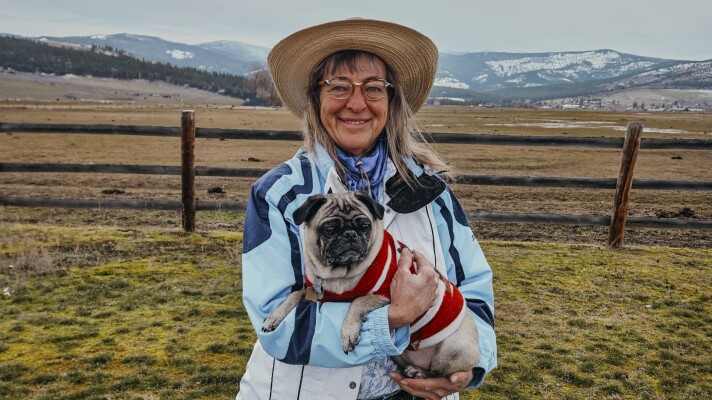 A woman wearing glasses, a wide-brimmed hat and a blue jacket smiles while holding a small pug dressed in a red sweater. They stand in a grassy field bordered by a wooden fence, with rolling hills and snow-dusted mountains in the background under a cloudy sky.