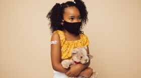 Small girl wearing protective face mask sitting with her teddy bear. Girl child with face mask on brown background after getting vaccination.