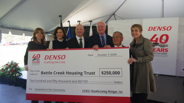 Three women and three men all hold up a large white check with red trim on the top and bottom. The amount is $250,000, paid to the Battle Creek Housing Trust, as written on the check. At the top right can be seen "DENSO," the company giving out the check, written in red letters. Below this reads their motto, "Crafting the Core." To the right of DENSO can be seen the words "40 years." The people holding the check are standing in front of a makeshift stage, the back of which also has DENSO's name written on it along with the 40 year anniversary.