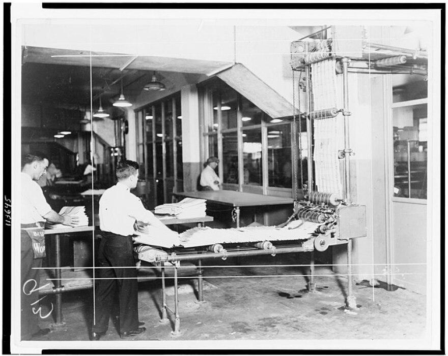 Interior view of newspaper delivery room showing man taking stack of newspapers from machine.