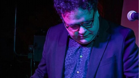 A man in a suitcoat and glasses stands at a mic while looking at a laptop and a mixing board