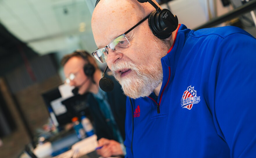 A bald man with a grey beard wears glasses, headphones and a bright blue fleece. He is standing next to another man wearing glasses and a headset.