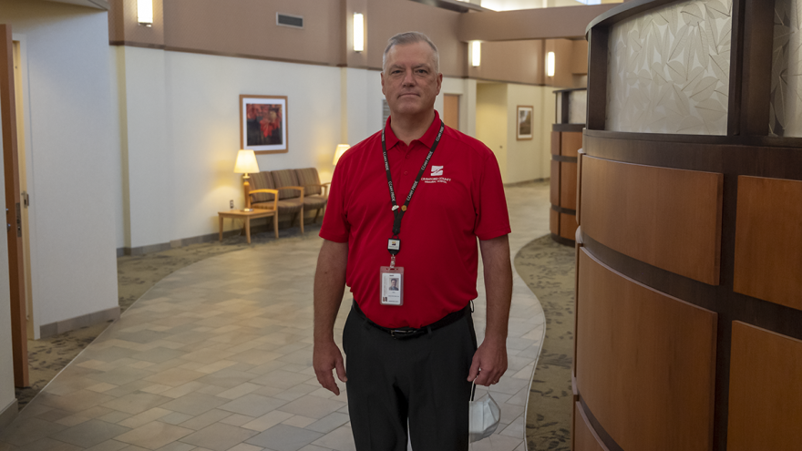 A man stands in a hall wearing a red polo shirt with a lanyard and ID card around his neck.