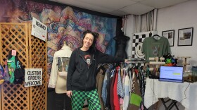 Person with green checkered pants and a black hoodie stands in a space with clothing on racks and hangers