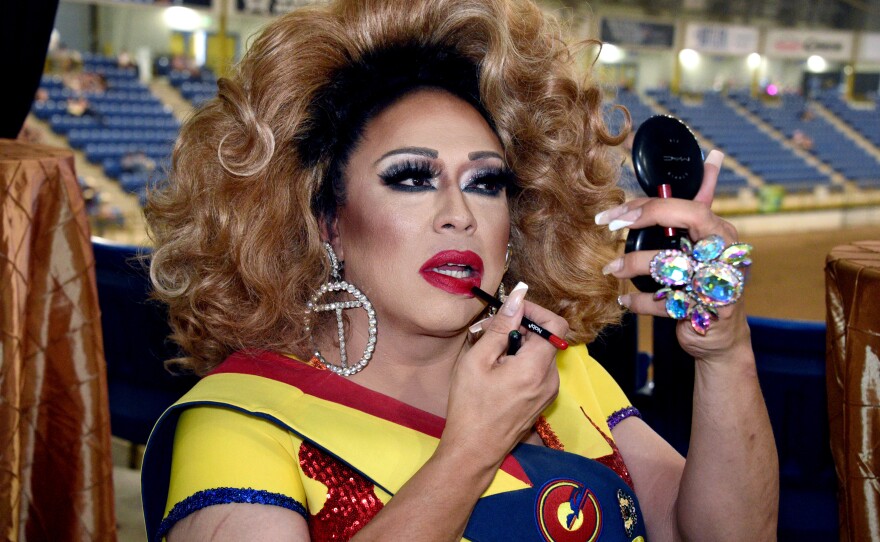 A drag queen puts on red lipstick while holding up a compact to see what she is doing in the mirror. She has large earrings in that are rhinestone circles with the letter "T" in the middle 